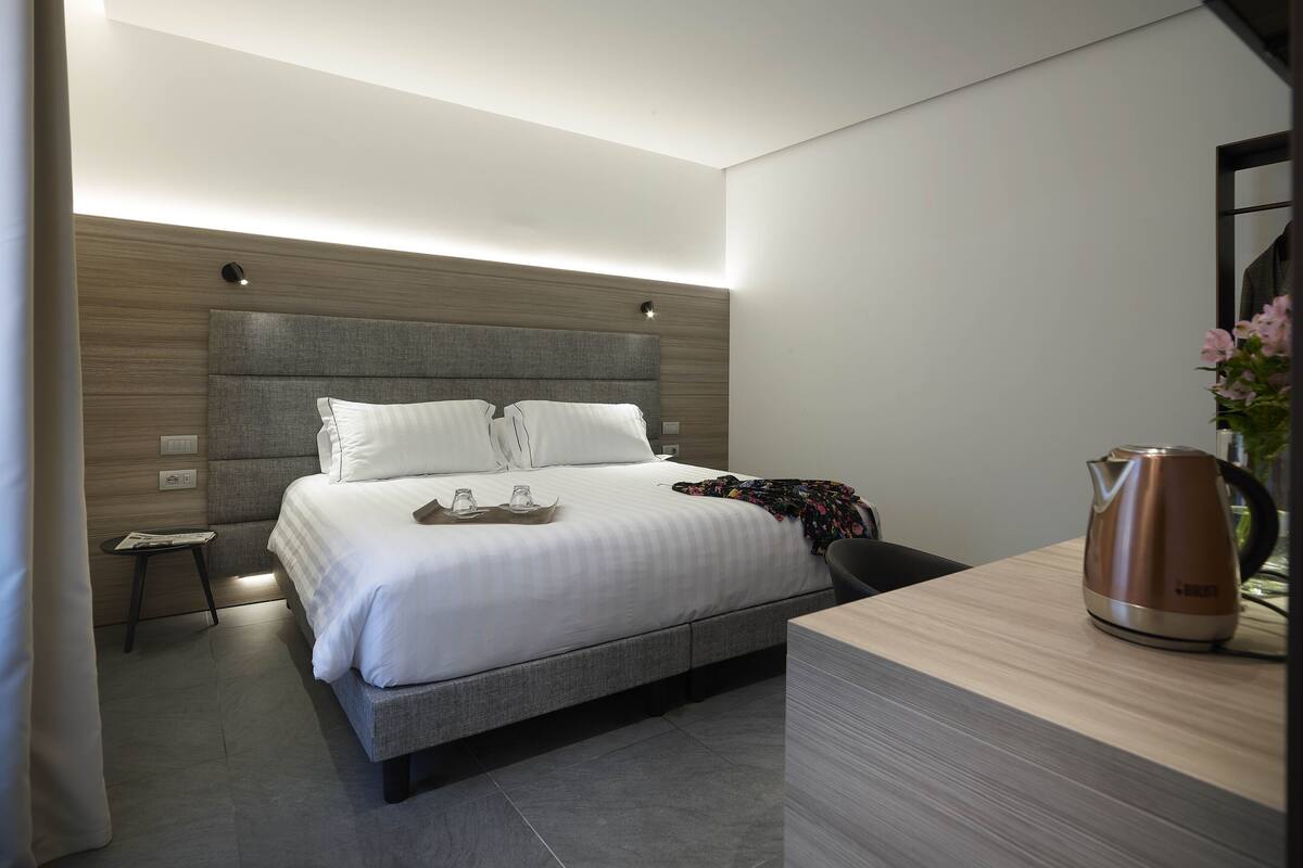 Ein modernes Hotelzimmer mit einem großen Bett, Nachttischen, einem Holz-Kopfteil und einer Kaffeemaschine auf dem Nachttisch.