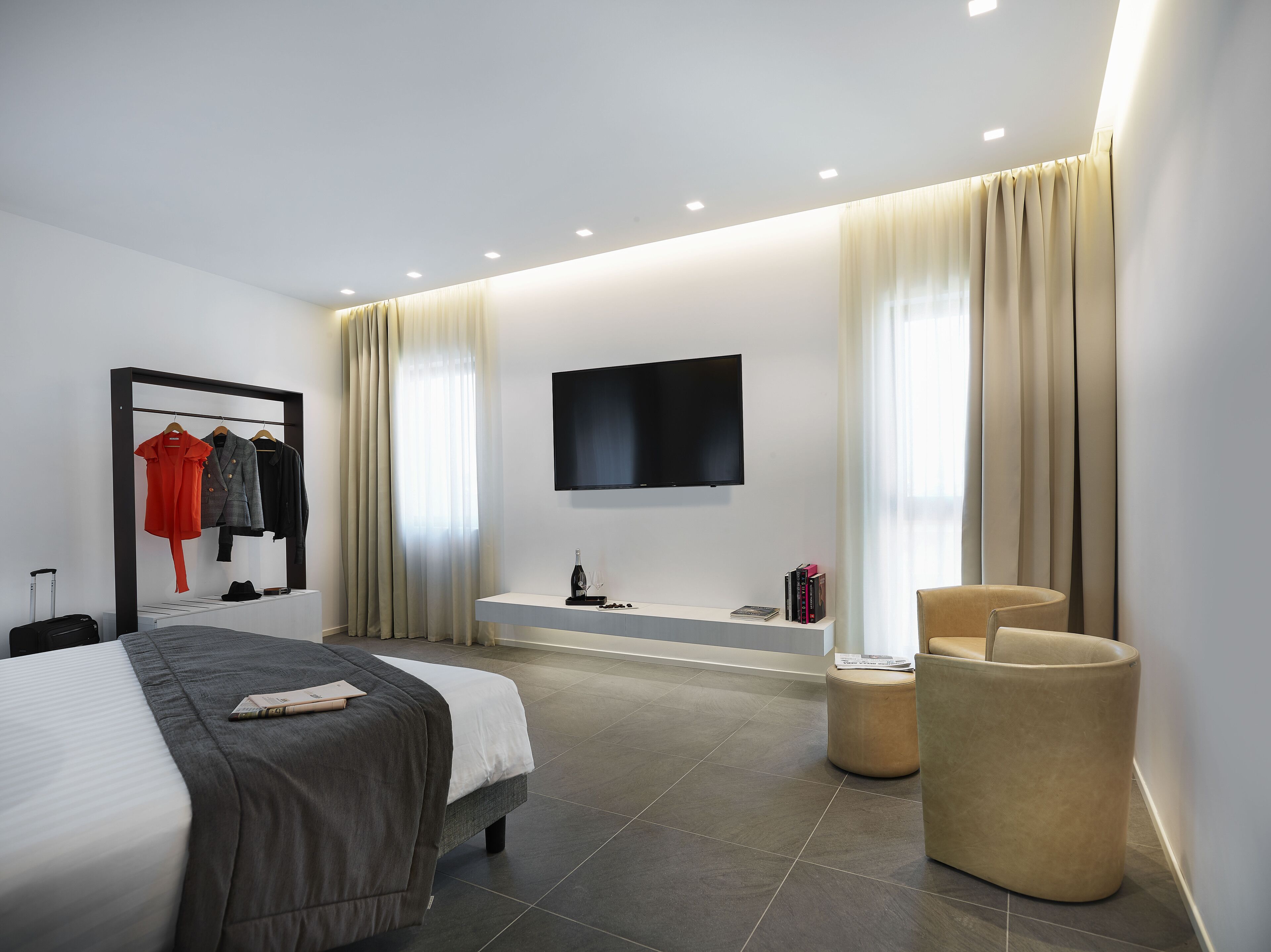 A modern hotel room with a bed, a seating area, a TV, and a clothing rack.