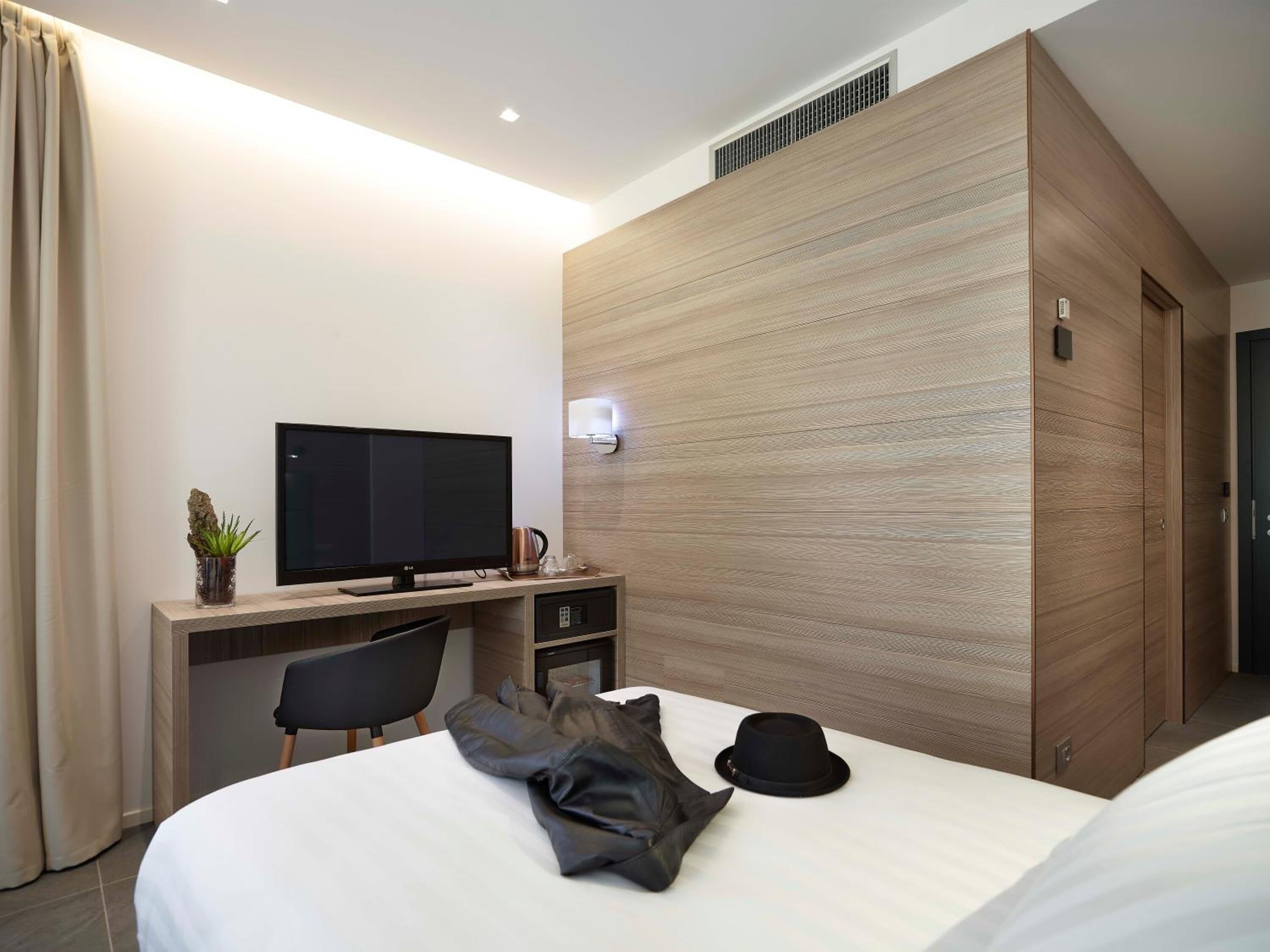 Quarto de hotel com uma cama, uma mesa com televisão, uma cafeteira e uma planta.