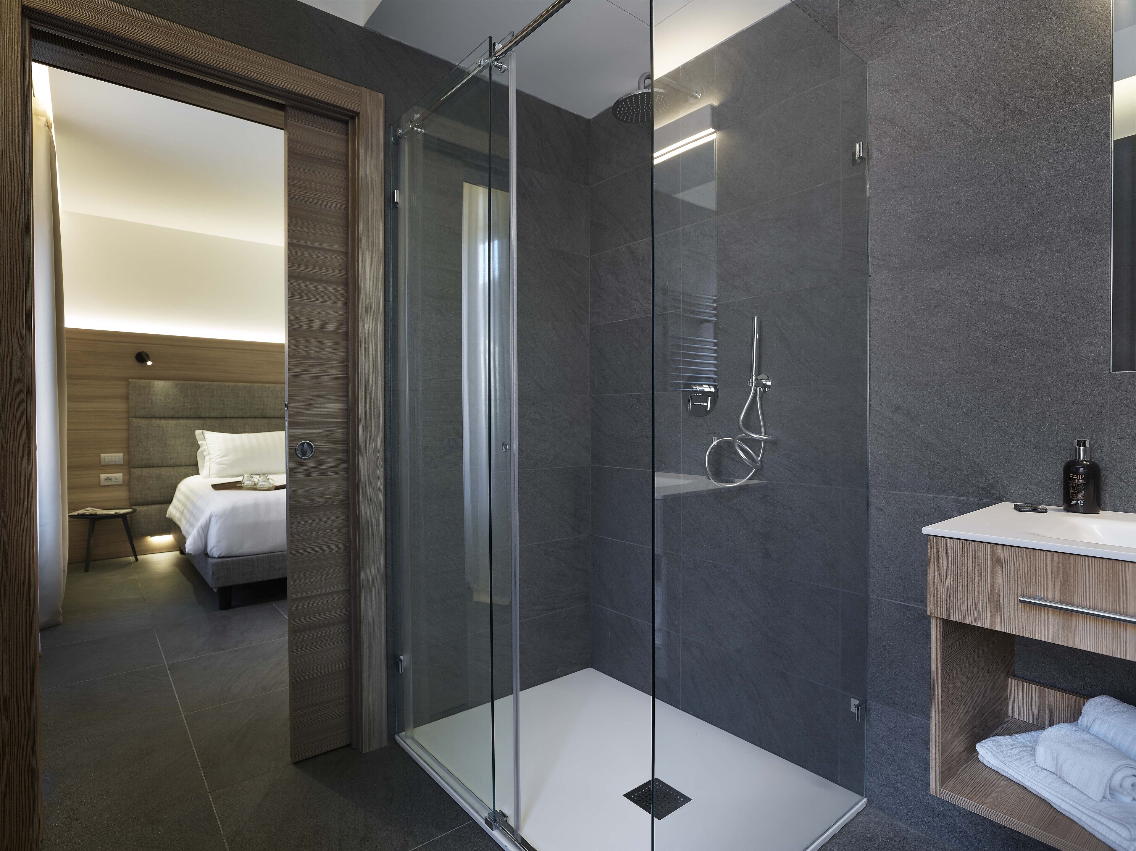 Une salle de bain moderne avec une douche à parois de verre, un meuble-lavabo en bois et un lit soigneusement fait dans la pièce adjacente.