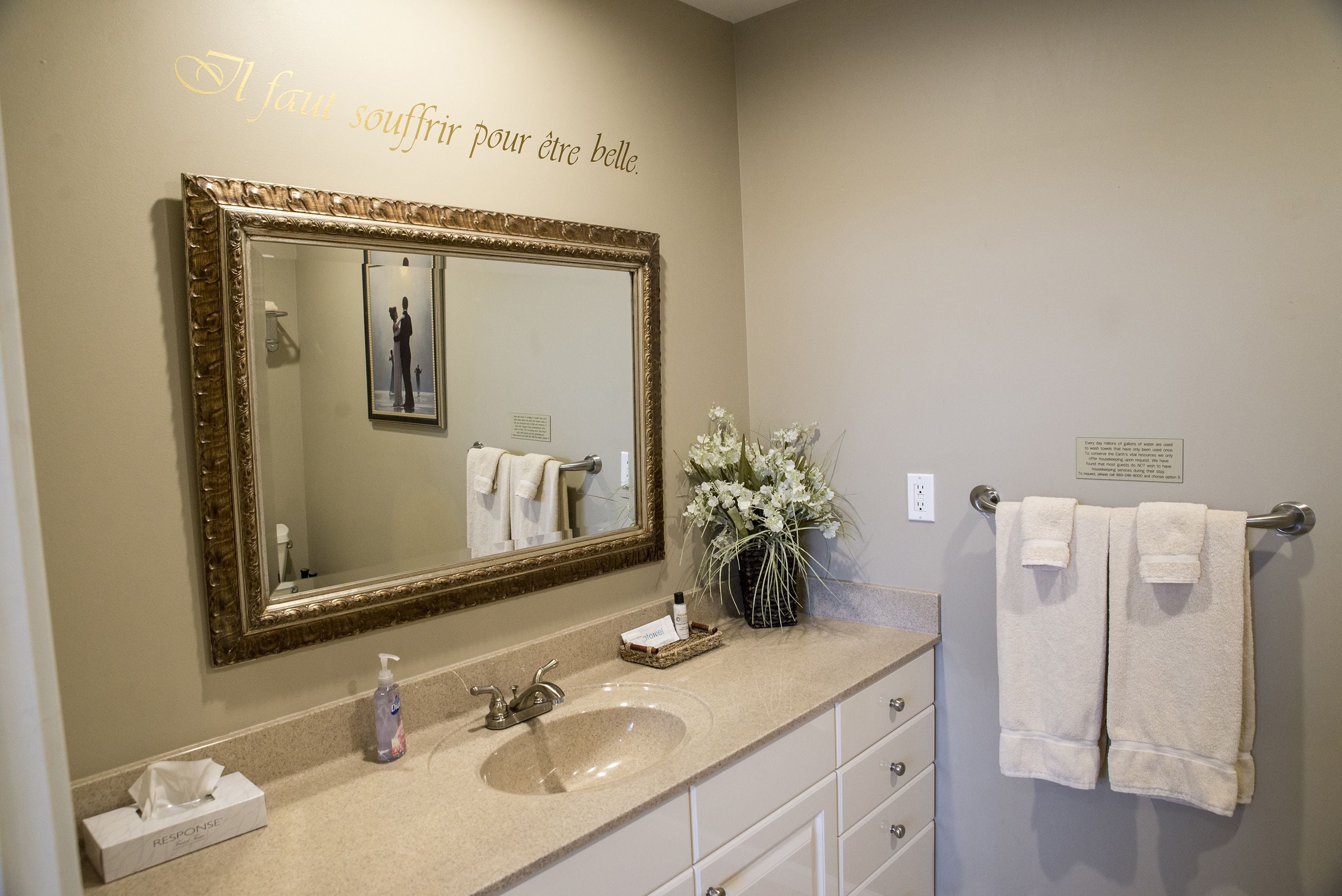 Un baño con un espejo grande, un lavabo, un perchero para toallas y un letrero decorativo en la pared.