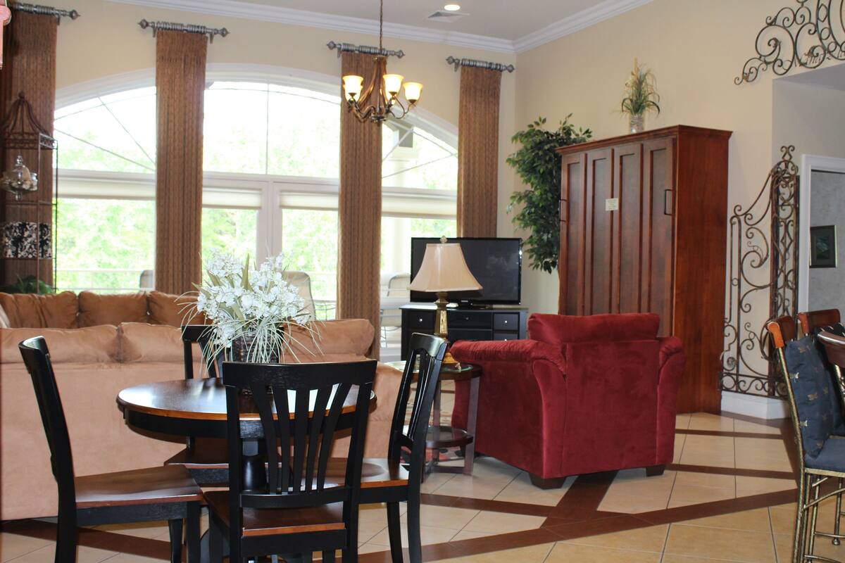A living room with a sofa, red armchair, dining table, and chairs. There is a television and a cabinet with a plant on top.