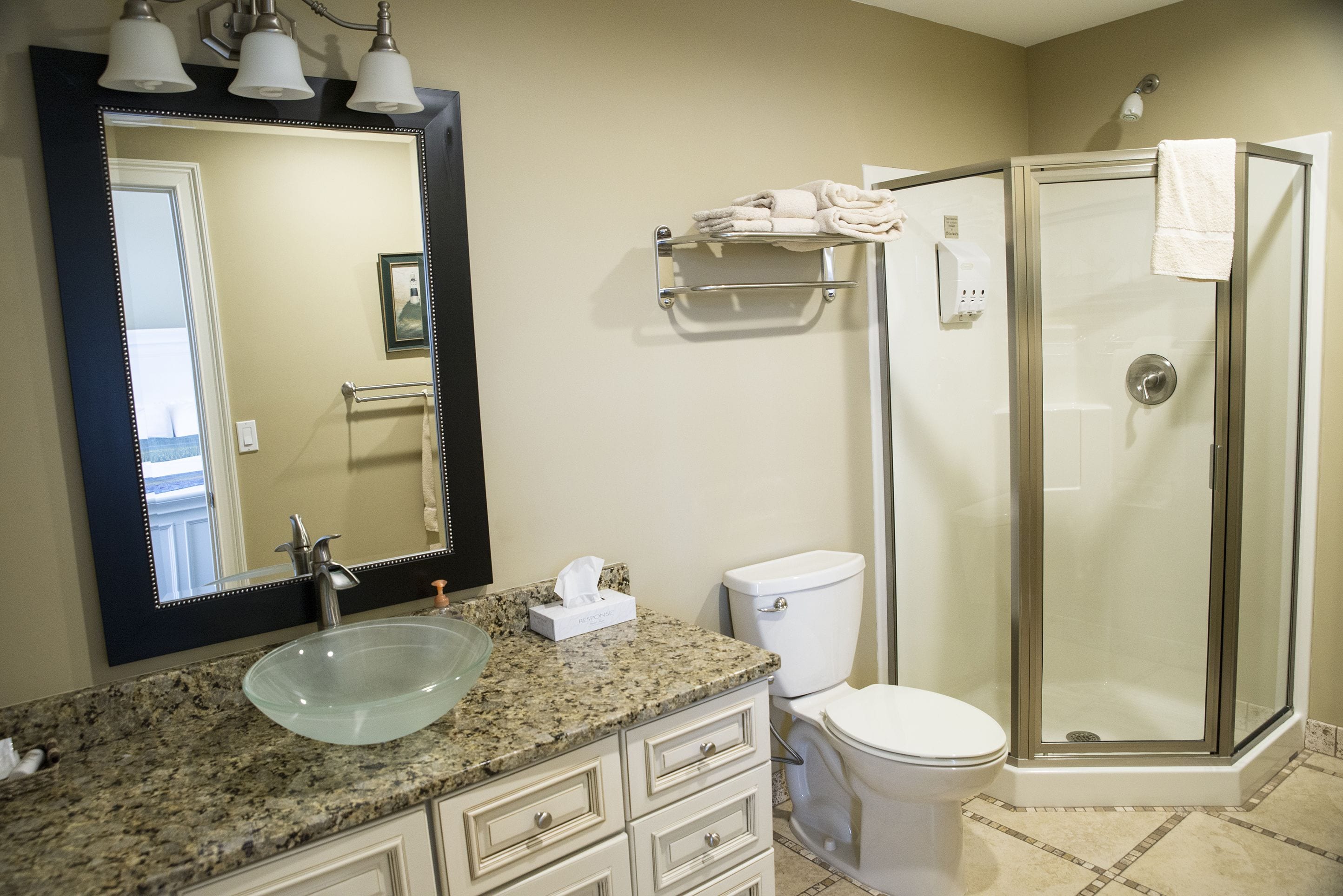 Une salle de bain dotée d’un comptoir en granit, d’un lavabo en verre, de toilettes blanches, d’une douche à parois de verre et d’un grand miroir avec trois lampes.