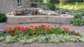 A stone building with a stone patio, a small waterfall feature, and a flower bed with red, purple, and white flowers. - Riverwalk Place Resort & Spa (Gladwin)