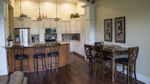 A kitchen with white cabinets, a central island with bar stools, a dining table with chairs, and pendant lighting. - Riverwalk Place Resort & Spa (Gladwin)