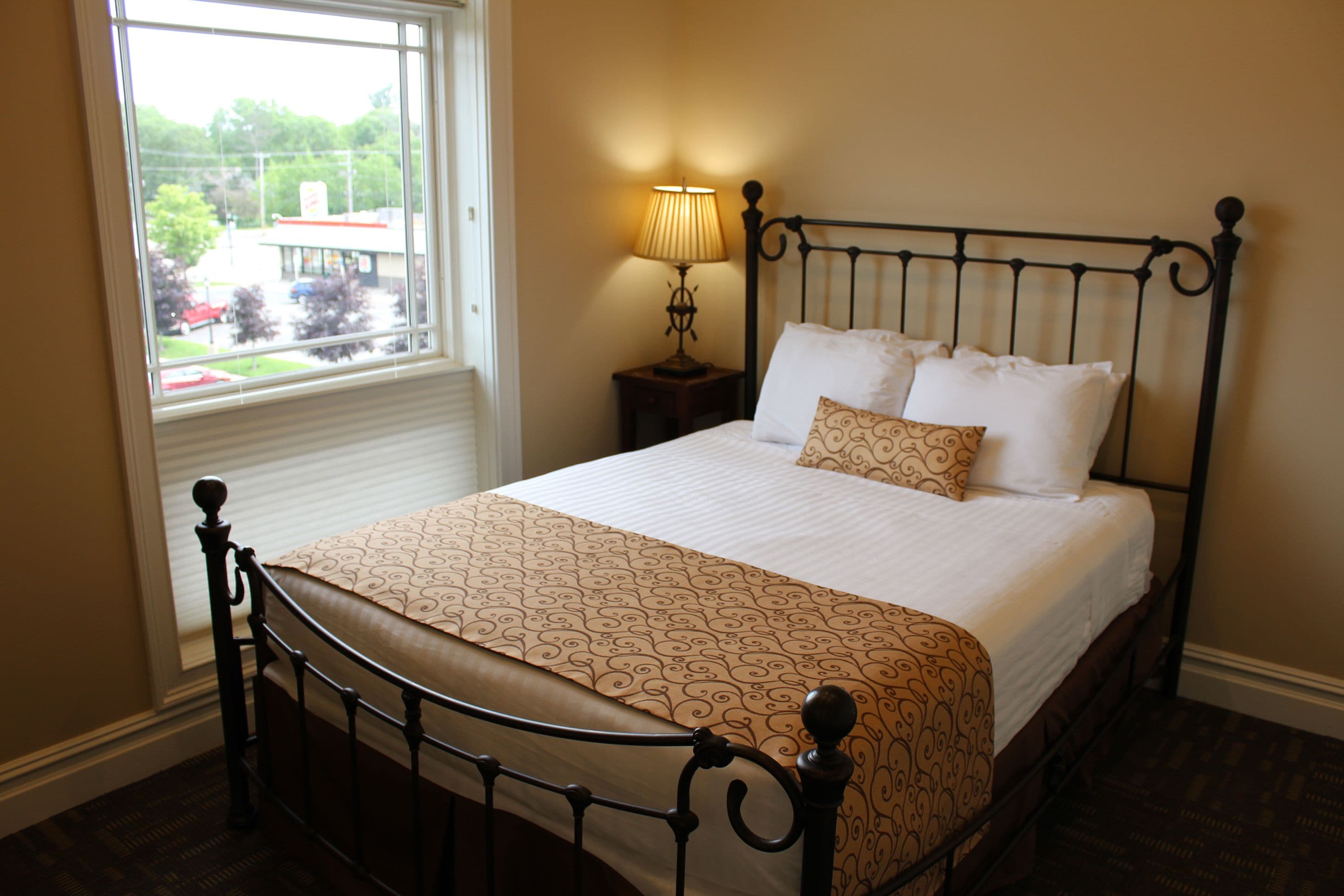 A neatly made bed with a patterned bedspread, a bedside lamp, and a window with a view of a building and trees.