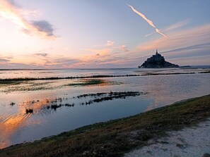 A coastal landscape with a prominent castle-like structure on a hill, surrounded by water and grassy areas.