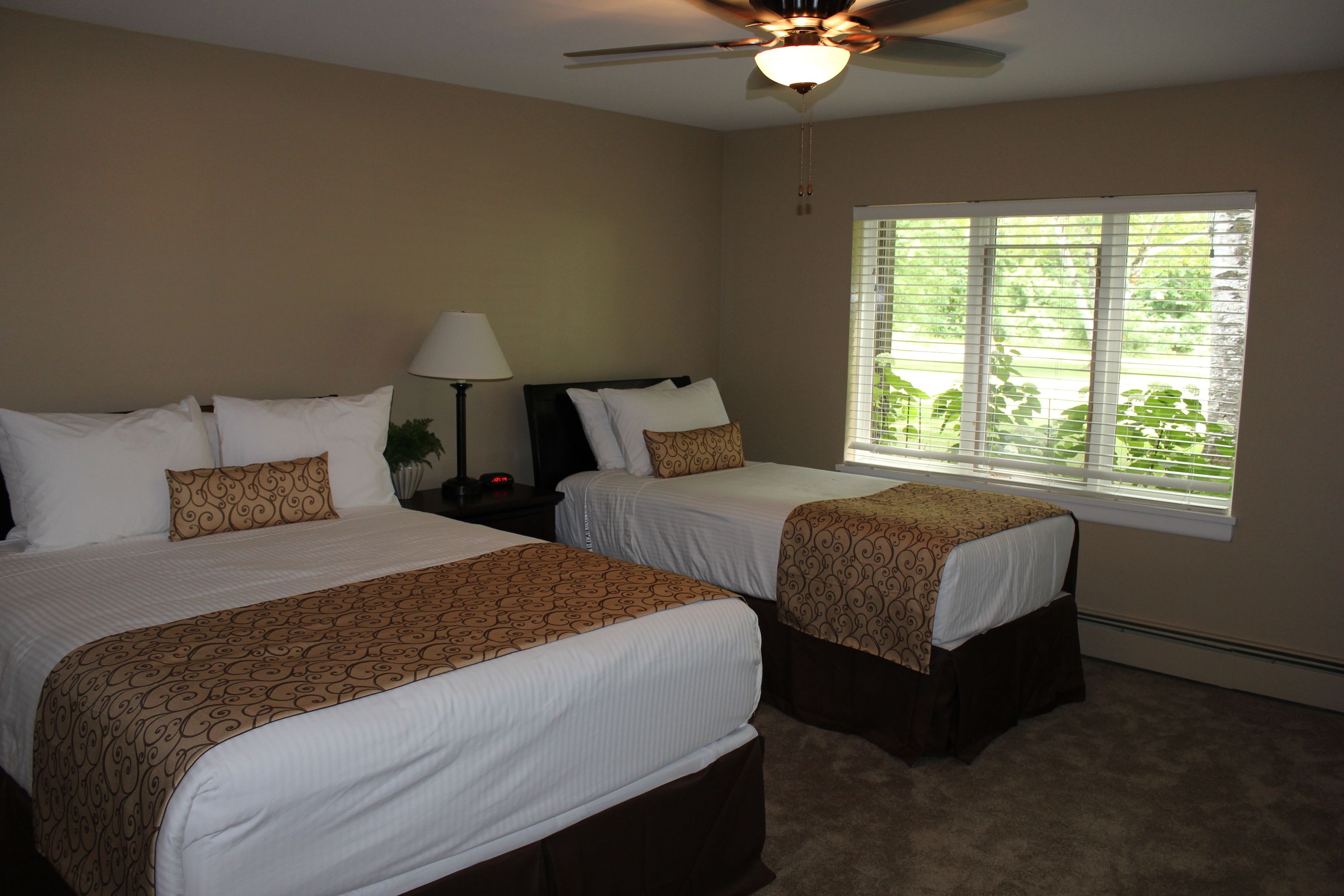 A hotel room with two beds, a ceiling fan, a window with blinds, and a nightstand with a lamp.