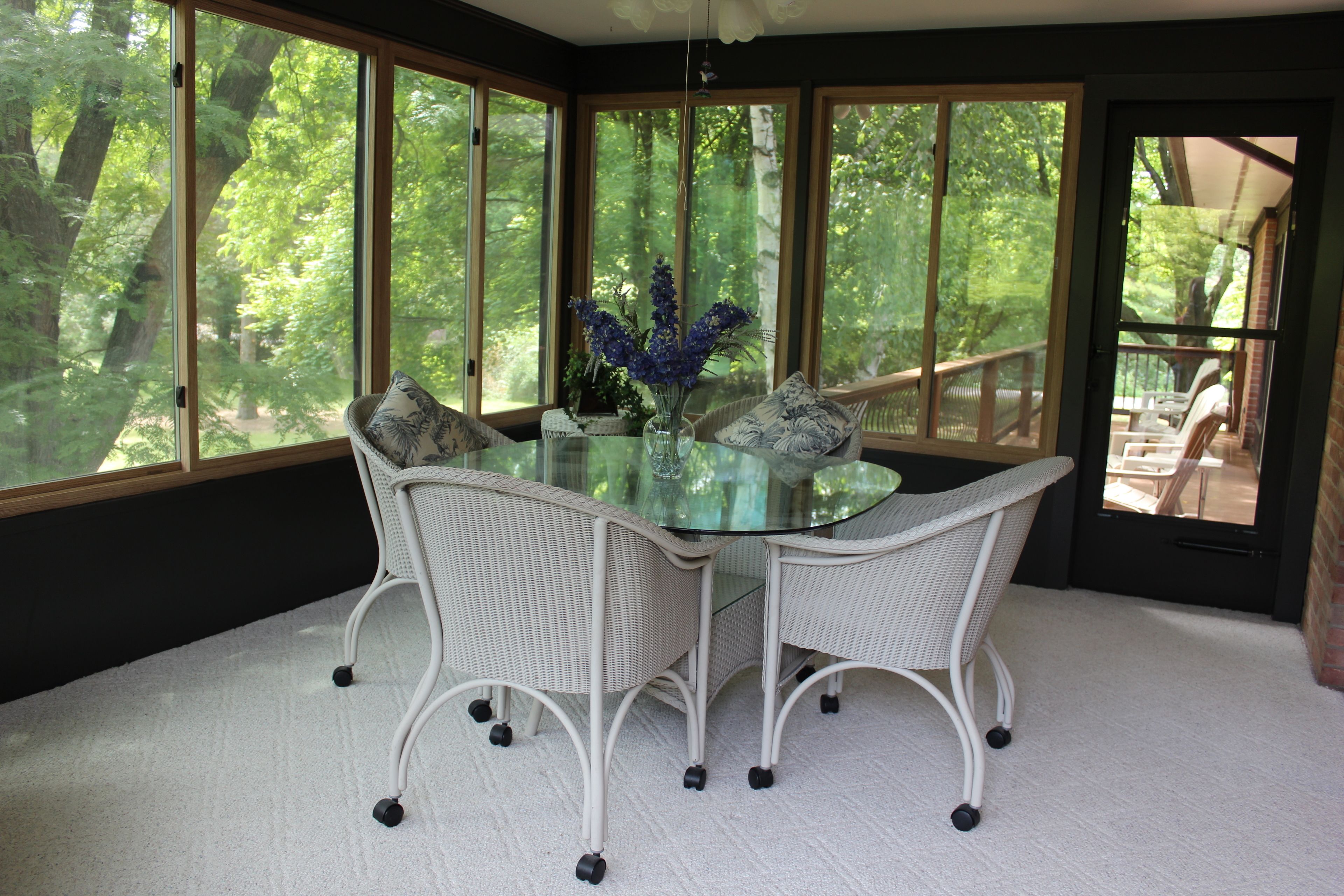 A dining area with a glass table surrounded by wicker chairs, a vase with flowers, and large windows offering a view of trees.