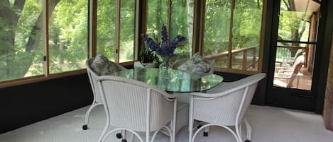A dining area with a glass table surrounded by wicker chairs, a vase with flowers, and large windows offering a view of trees.