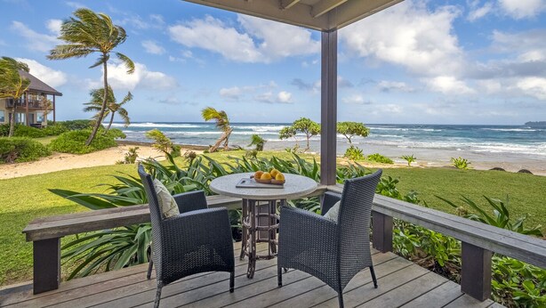 Uma área de estar coberta ao ar livre, com mesa e cadeiras, com vista para uma praia com palmeiras e ondas do oceano.