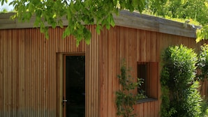 Une cabane en bois avec une porte en verre, une petite fenêtre et une terrasse en bois donnant sur un étang.