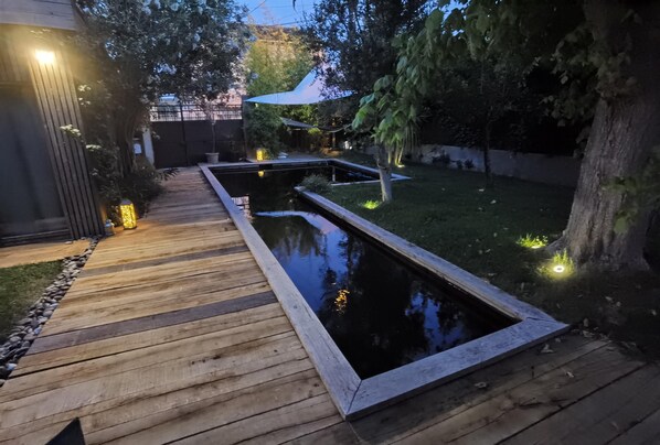 Eine Holzterrasse führt zu einem rechteckigen Teich mit Steinrand, umgeben von Gras und Bäumen.