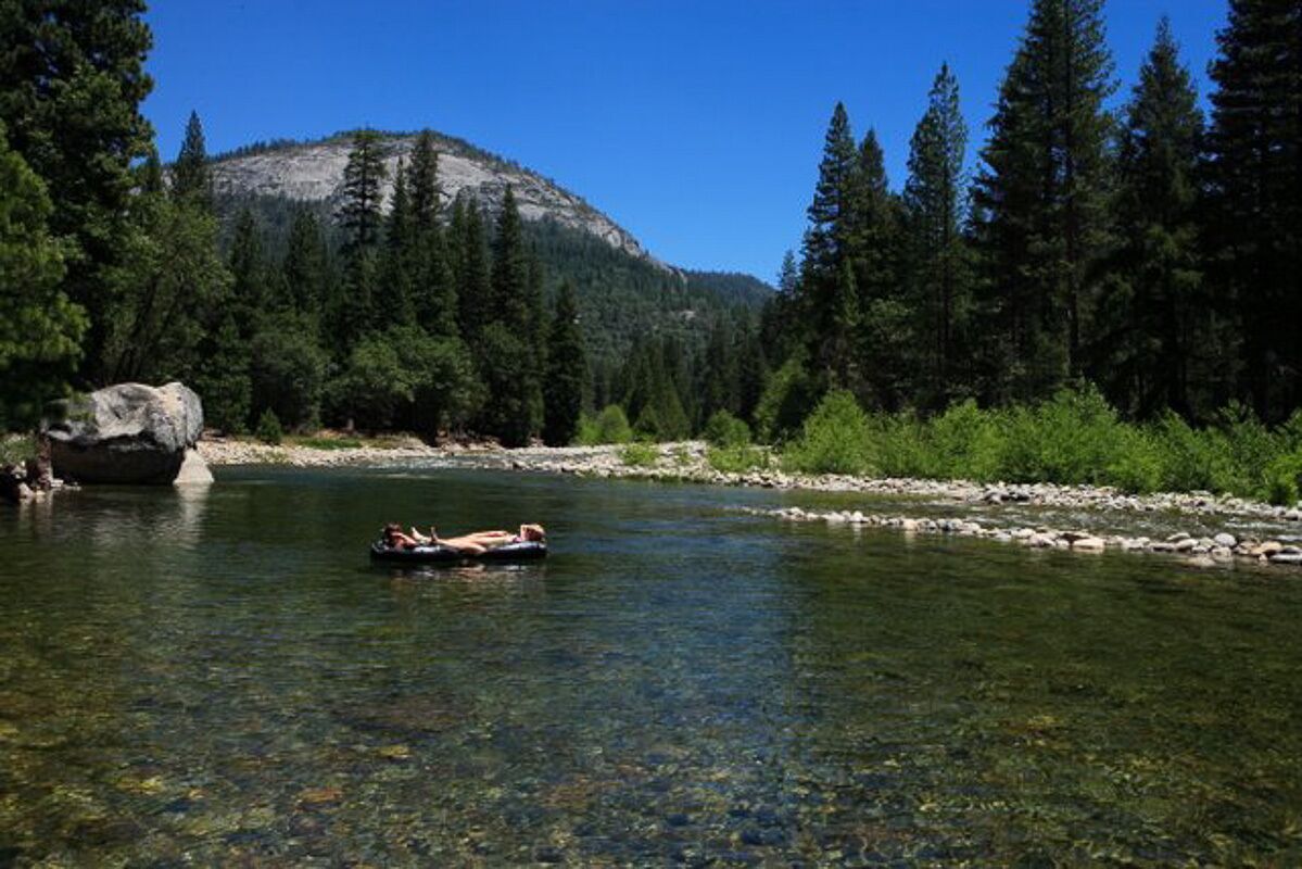 Un río claro con una persona flotando en una balsa, rodeada de un bosque denso y una montaña al fondo.