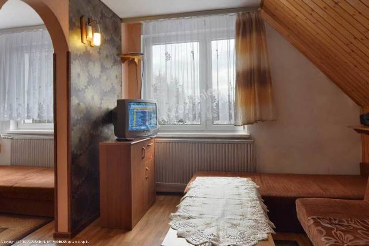 A small, cozy room with a wooden ceiling, a window with curtains, a television on a wooden stand, and a bed with a patterned bedspread.