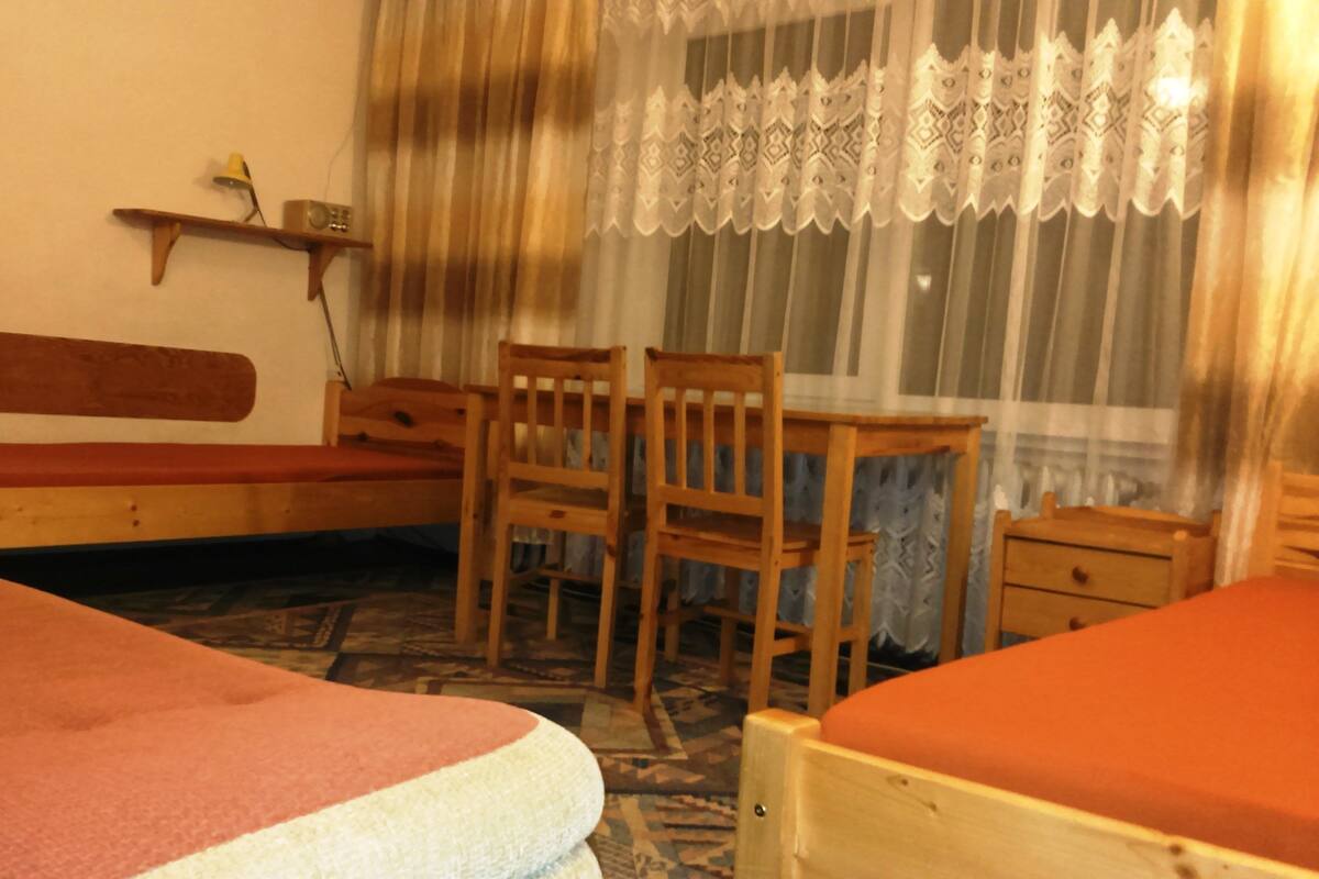 A room with a bed, a desk, and a chair. There is a shelf with a lamp and a radio.