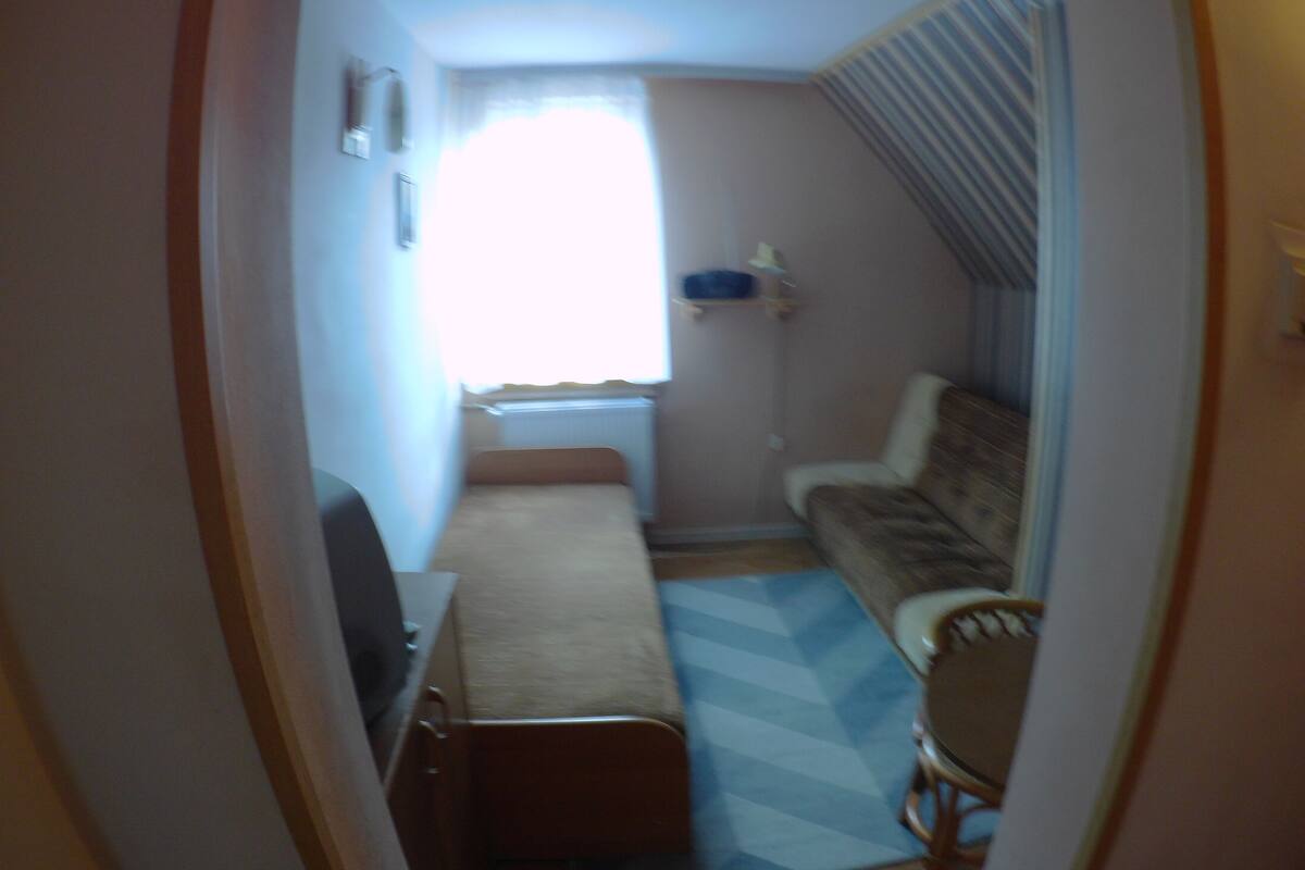 A narrow room with a bed, a chair, a small table, and a window with curtains.