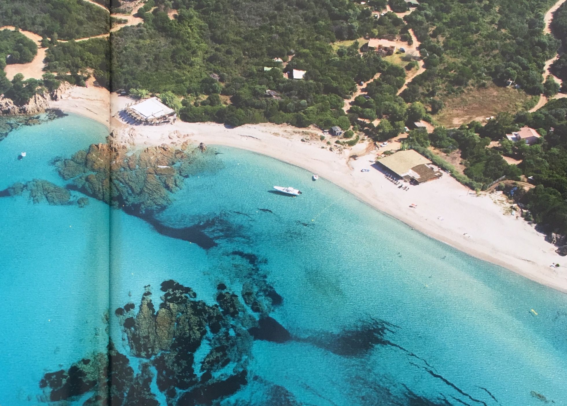 Vista aérea de uma área costeira com praia arenosa, águas azul-claras, afloramentos rochosos e edifícios dispersos.