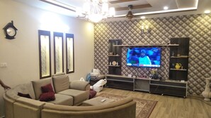 A living room with a beige sofa, a flat-screen TV mounted on a textured wall, a chandelier, and a wooden ceiling.