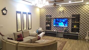 A living room with a beige sofa, a flat-screen TV mounted on a textured wall, a chandelier, and a wooden ceiling.
