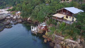 A coastal house with a balcony, surrounded by lush greenery and a rocky shoreline.