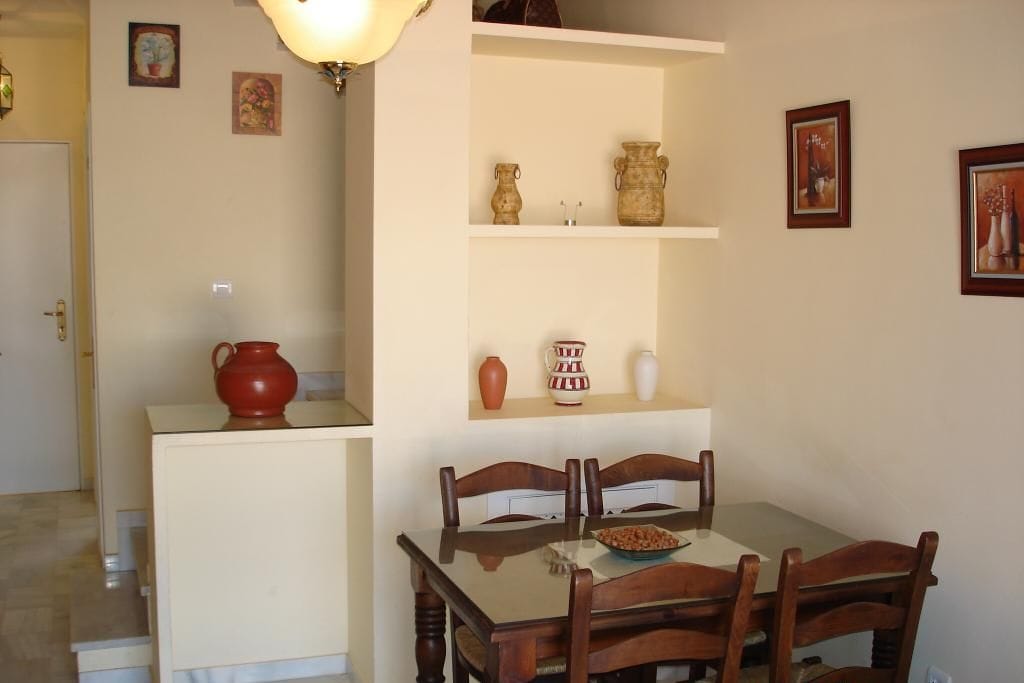 A dining area with a wooden table and chairs, a small kitchen counter with a red pot, and shelves with decorative items.