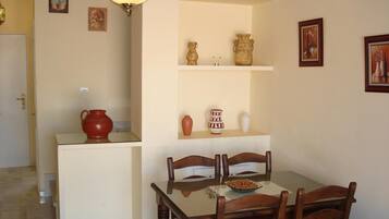 A dining area with a wooden table and chairs, a small kitchen counter with a red pot, and shelves with decorative items.