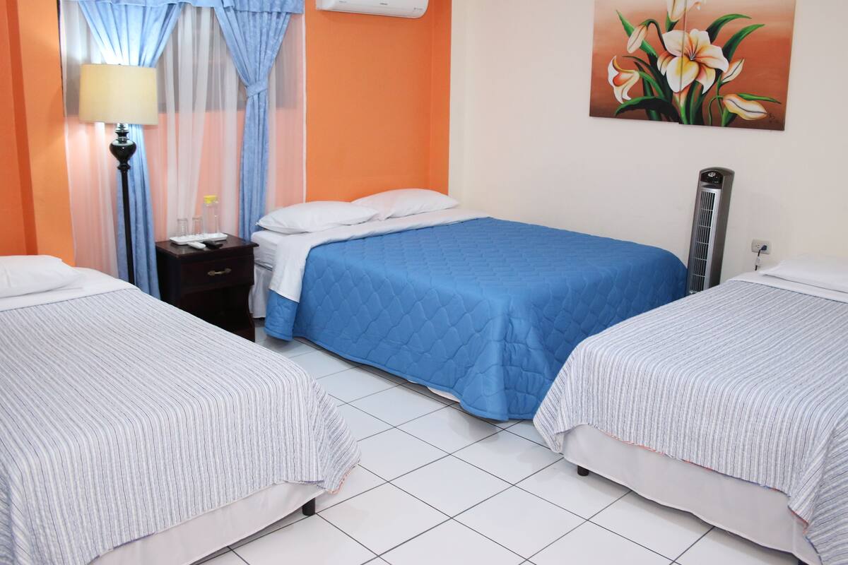 A hotel room with two single beds, a nightstand, an air conditioning unit, a lamp, and a painting on the wall.