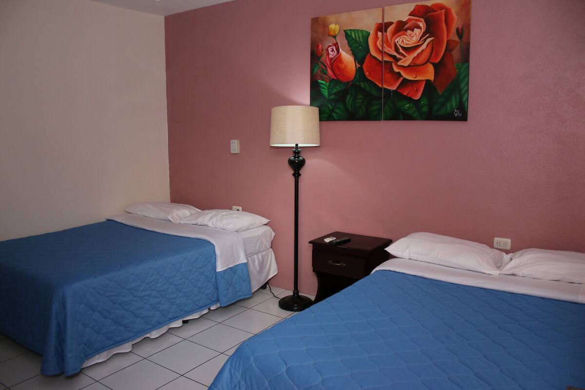 A room with two beds, each with blue bedding and white pillows, a nightstand with a lamp, and a painting of roses on the wall.