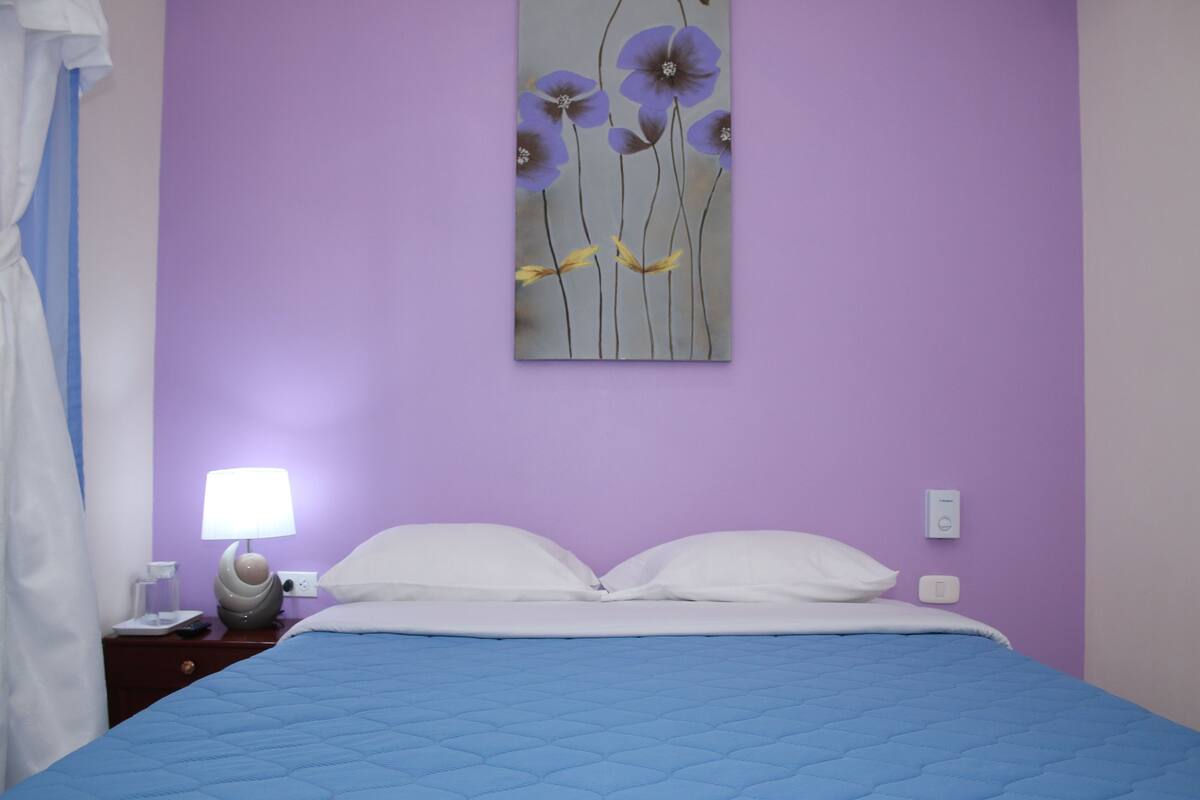 A bedroom with a blue quilted bedspread, white pillows, a nightstand with a lamp, and a wall-mounted painting of flowers.