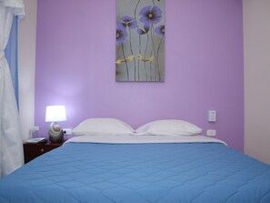 Un dormitorio con un edredón azul acolchado, almohadas blancas, una mesita de noche con lámpara y un cuadro de flores colgado en la pared.