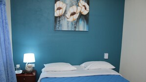 A bedroom with a blue bedspread, white pillows, a bedside lamp, and a painting of flowers on the wall. - Brisas Hotel (Matagalpa)