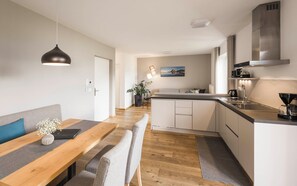 A modern kitchen with a central island, a dining area with a wooden table and bench, and a living space with a sofa and a painting on the wall.