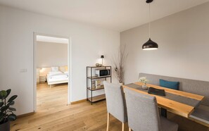 A modern, minimalistic room with a wooden dining table, grey upholstered chairs, a grey sofa, a black pendant light, and a white bed in the background.