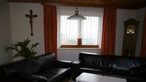 A living room with a black leather sofa, a white coffee table, a potted plant, a wooden cross on the wall, and a clock. - Apartment for up to 6 people, beautiful view (Hunderdorf)