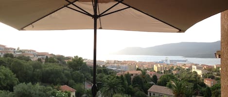 A balcony with a large umbrella, overlooking a coastal town with buildings and greenery.