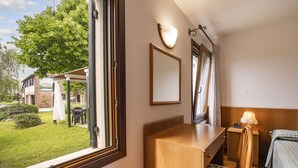 A hotel room with a wooden desk, a bed with a green striped bedspread, a nightstand with a lamp, a window with a view of the garden and building outside, and a mirror on the wall. - 2 bedroom apartment, Mira, Venice, with swimming pool (Oriago)