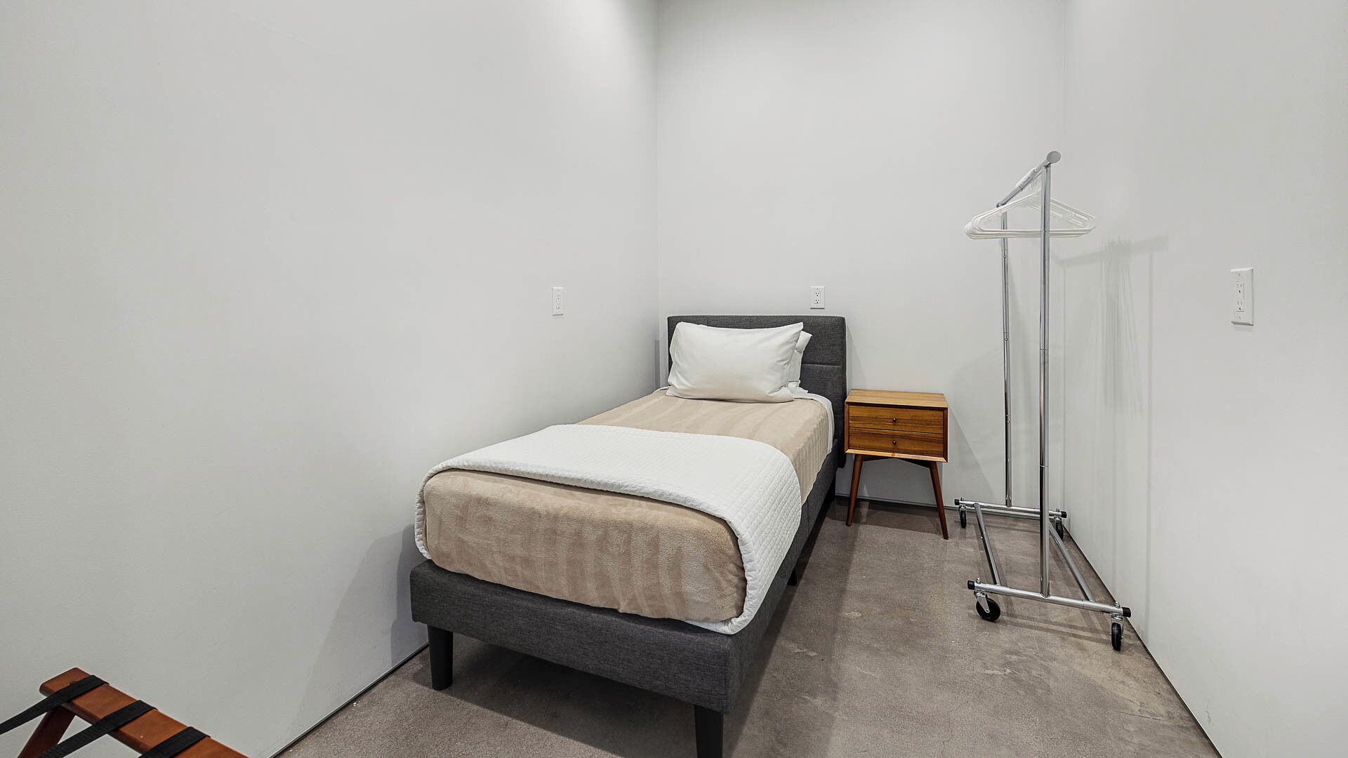 A small, neatly arranged bedroom with a bed, bedside table, and a clothes drying rack.