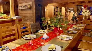 Interior de uma cabana de madeira com uma longa mesa de jantar posta para uma refeição, decorada com árvores de Natal e uma toalha de mesa vermelha.