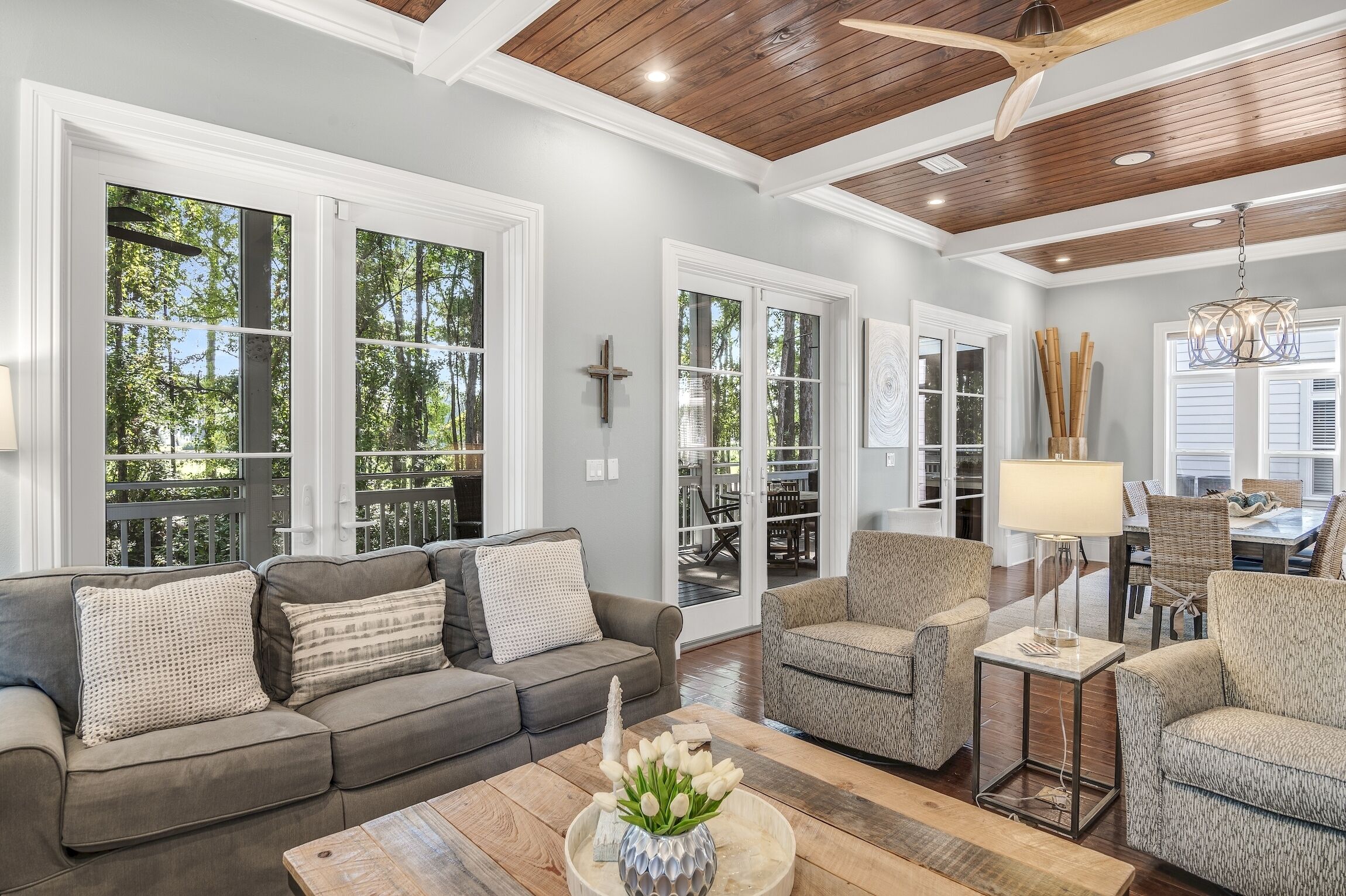 A spacious living room with a gray sofa, two armchairs, and a coffee table. Large windows provide natural light and a view of the outdoors.