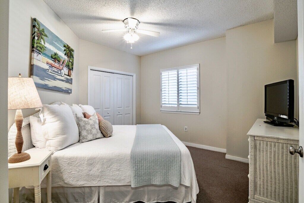Une chambre à coucher comprenant un lit, une table de chevet avec une lampe, un téléviseur posé sur un meuble, un ventilateur de plafond, une porte et une fenêtre avec des stores.