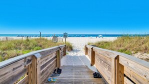 Ein hölzerner Steg führt zu einem Strand mit Sonnenschirmen und blauem Himmel.