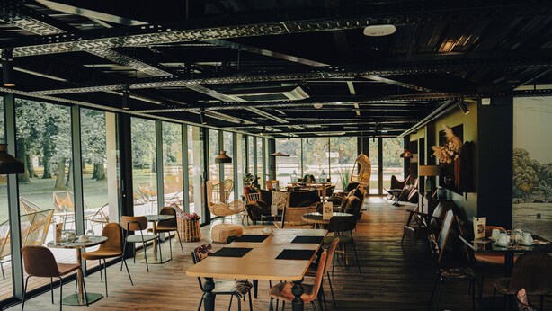 A spacious indoor area with multiple tables and chairs, large windows offering views of the outdoors, and a modern ceiling design. - Hôtel Le Corbusson Laval Ouest (Saint-Berthevin)