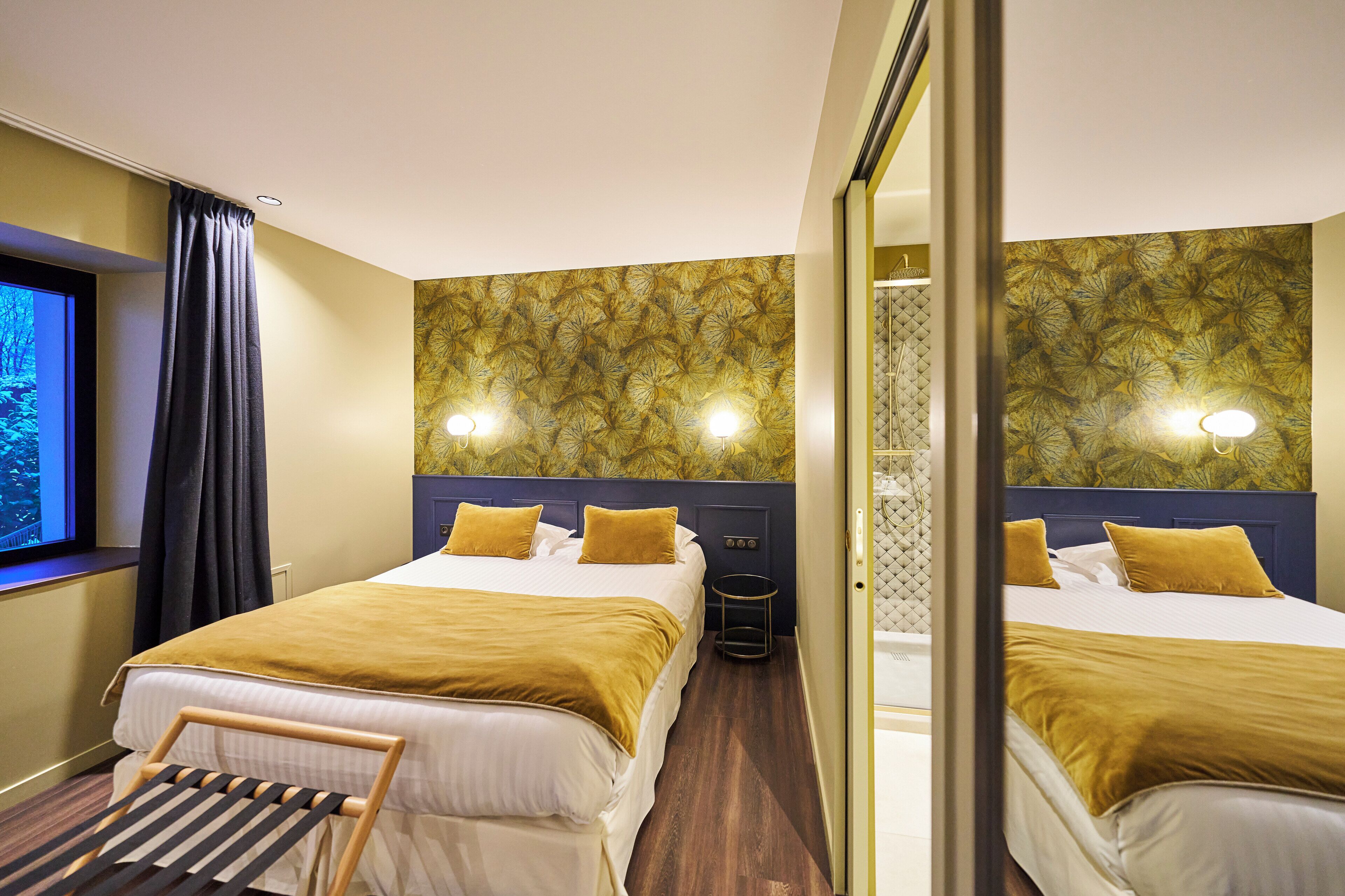 Habitación de hotel con dos camas, una ventana con cortinas, un espejo y un papel tapiz estampado.