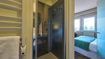 A modern bathroom with a glass shower enclosure, a white towel rack, and a visible window in the adjacent bedroom.