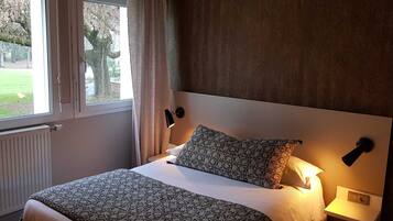 Una cama bien hecha con ropa de cama estampada, una lámpara de noche y vistas a un árbol fuera de la ventana.