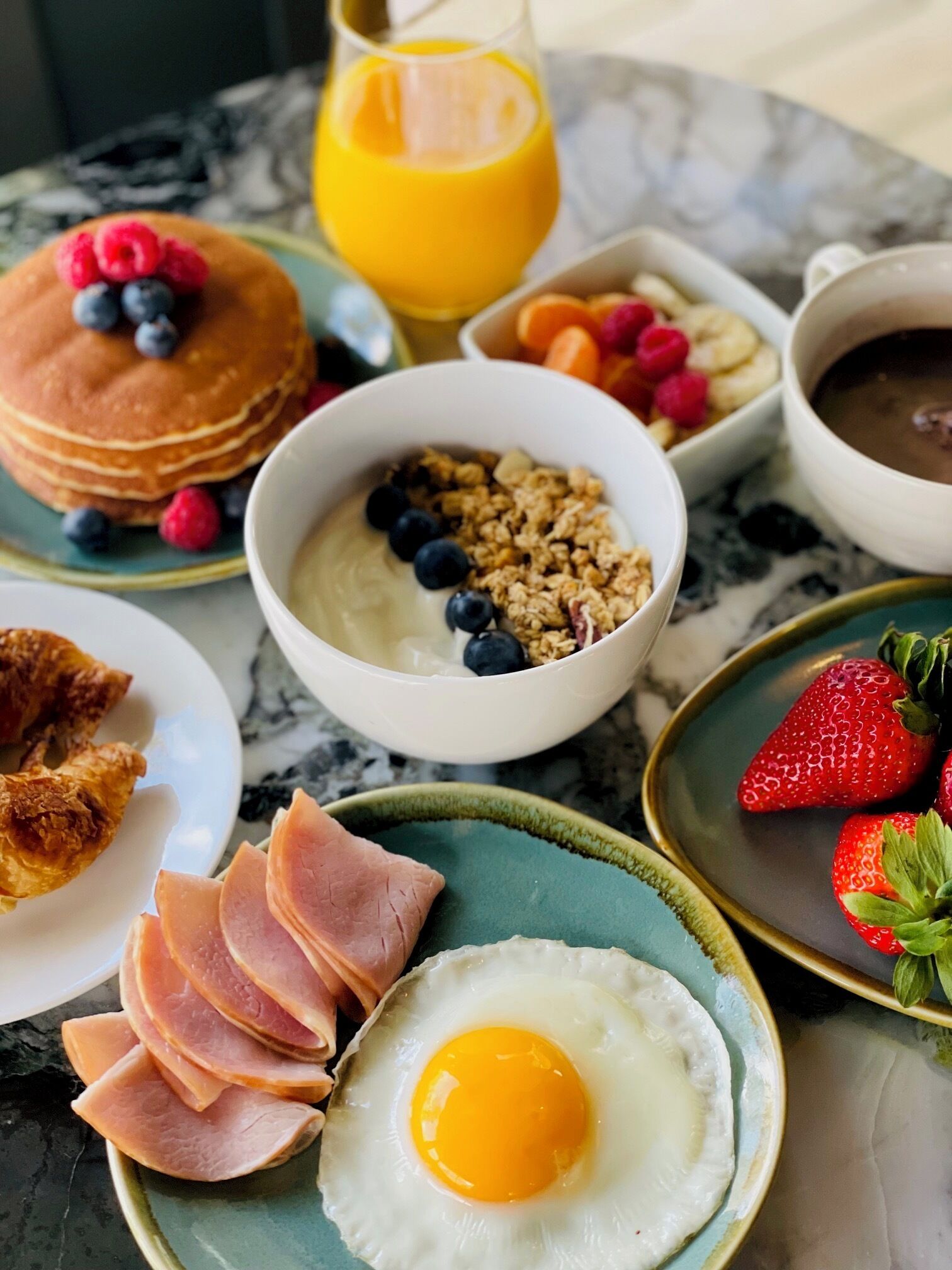 Una mesa de mármol con un desayuno que incluye panqueques, huevos revueltos, jamón, fresas y un tazón de yogur con frutos rojos.