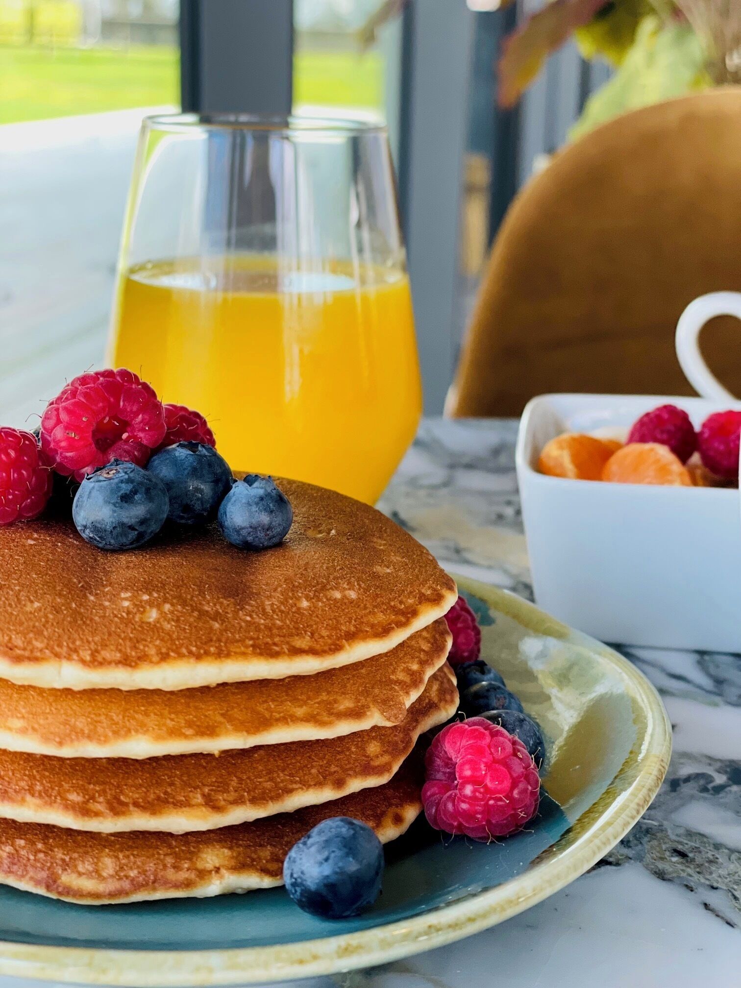 Una pila de tortitas con arándanos y frambuesas en un plato, un vaso de zumo de naranja y un bol con más bayas.