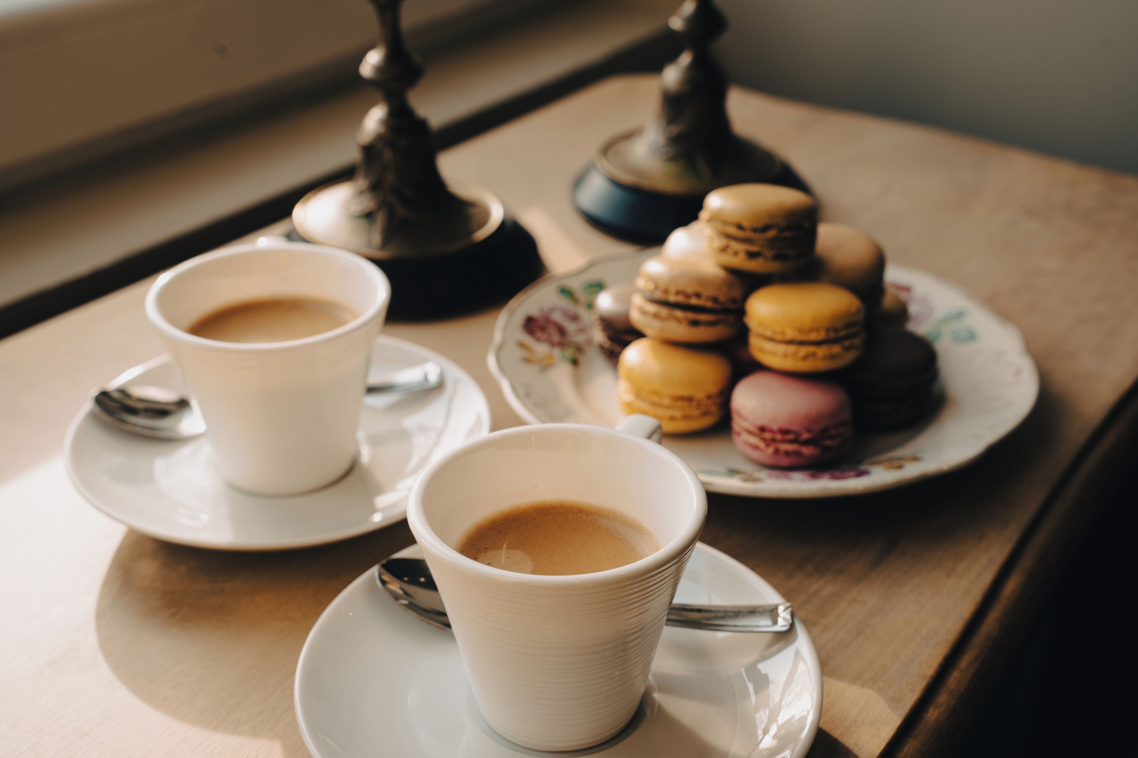 Two cups of coffee on a table with a plate of macarons and a spoon.