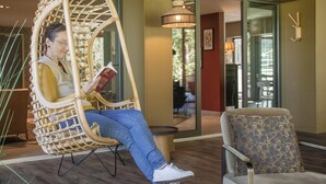 A person seated in a wicker chair reading a book in a room with a modern interior, a small round table, and a patterned armchair. - Hôtel Le Corbusson Laval Ouest (Saint-Berthevin)